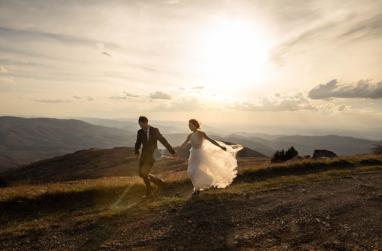 Photo of a bride and groom running across a mountaintop at sunset with sun flare. Whitetop Mountain, southwest Virginia