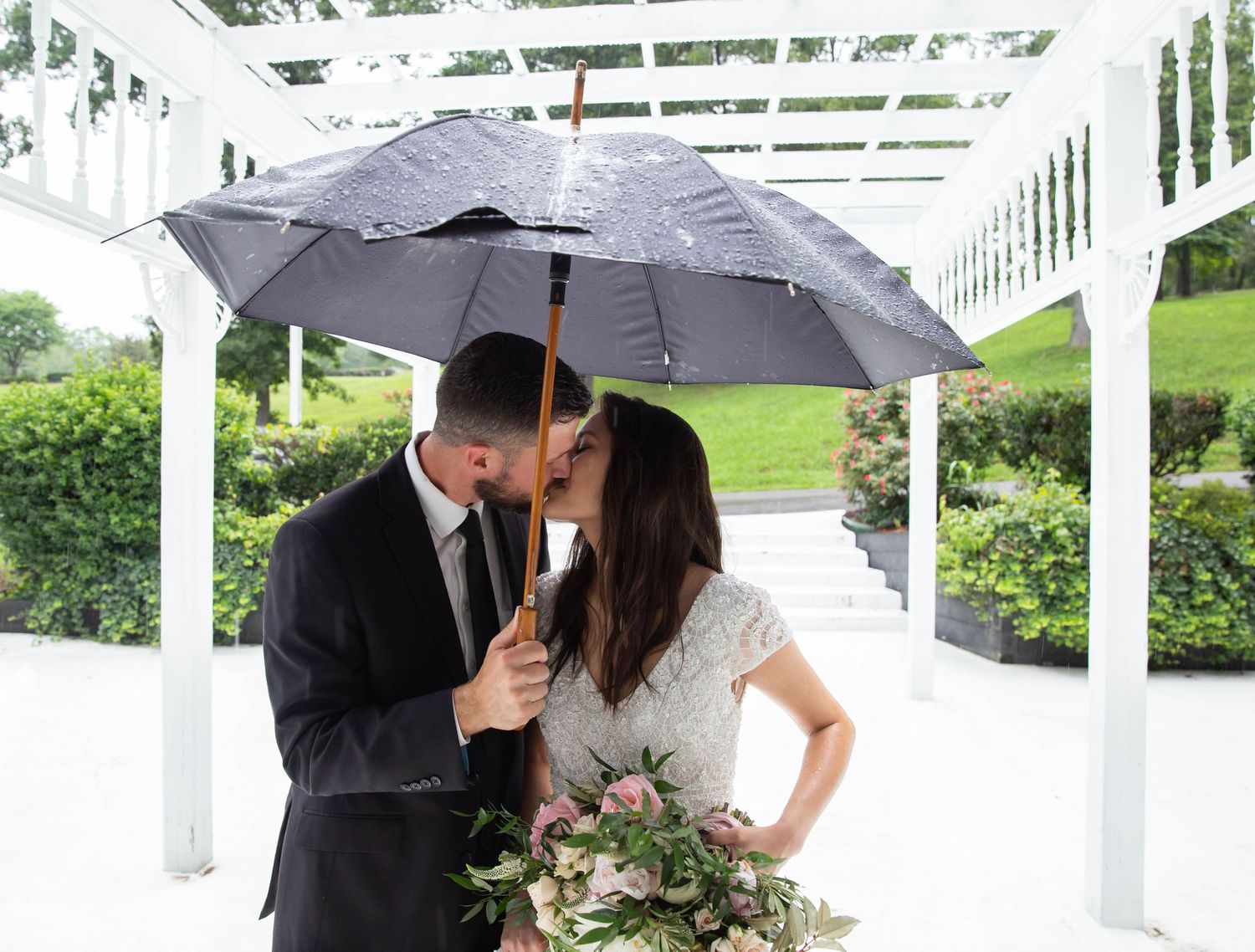 Photo of a bride and groom kissing while standing under a black umbrella in the raining