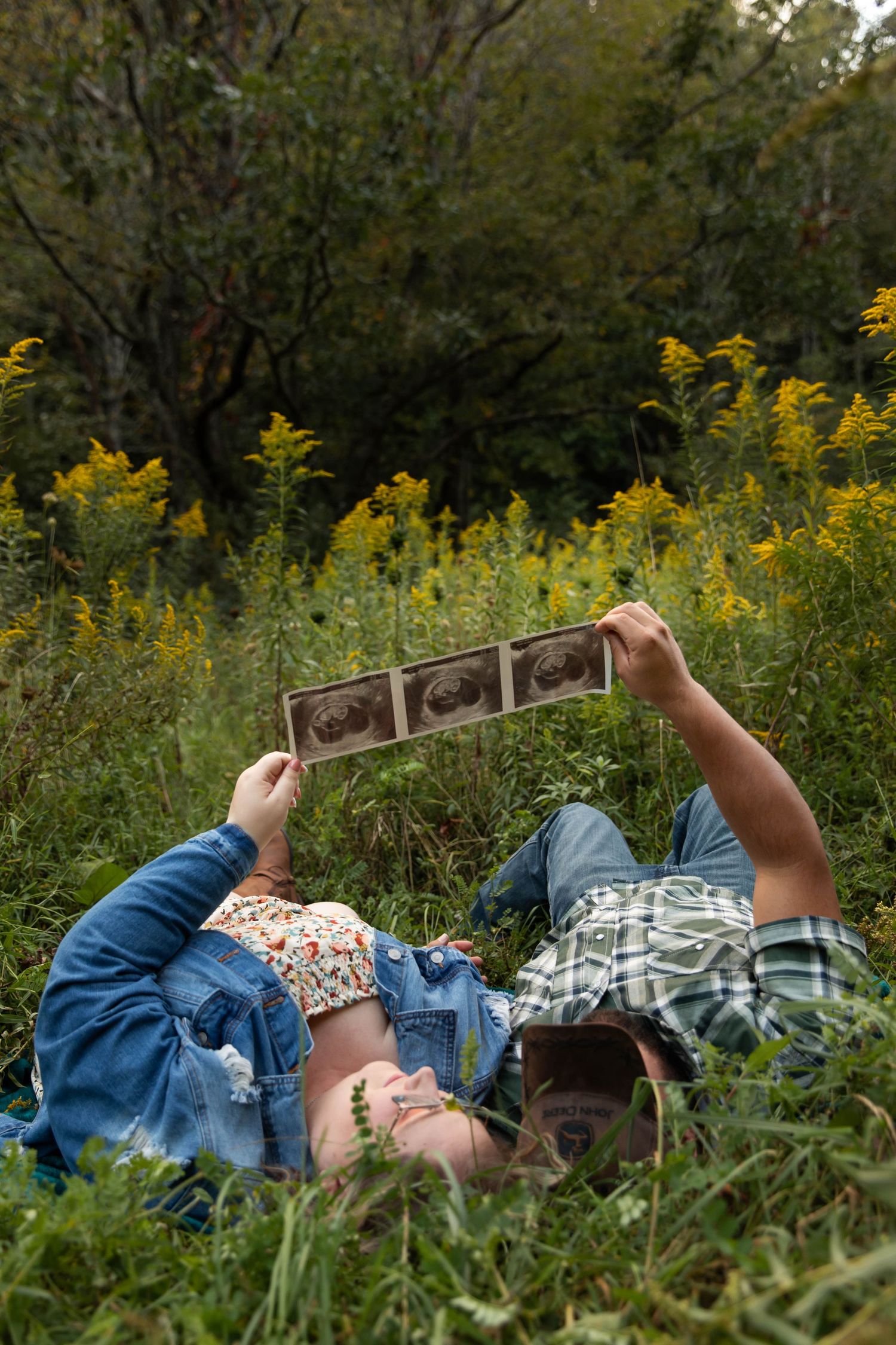 Photo of a couple laying in a field of yellow flowers , reading books that say "Mom and Dad" for a pregnancy announcement photo shoot