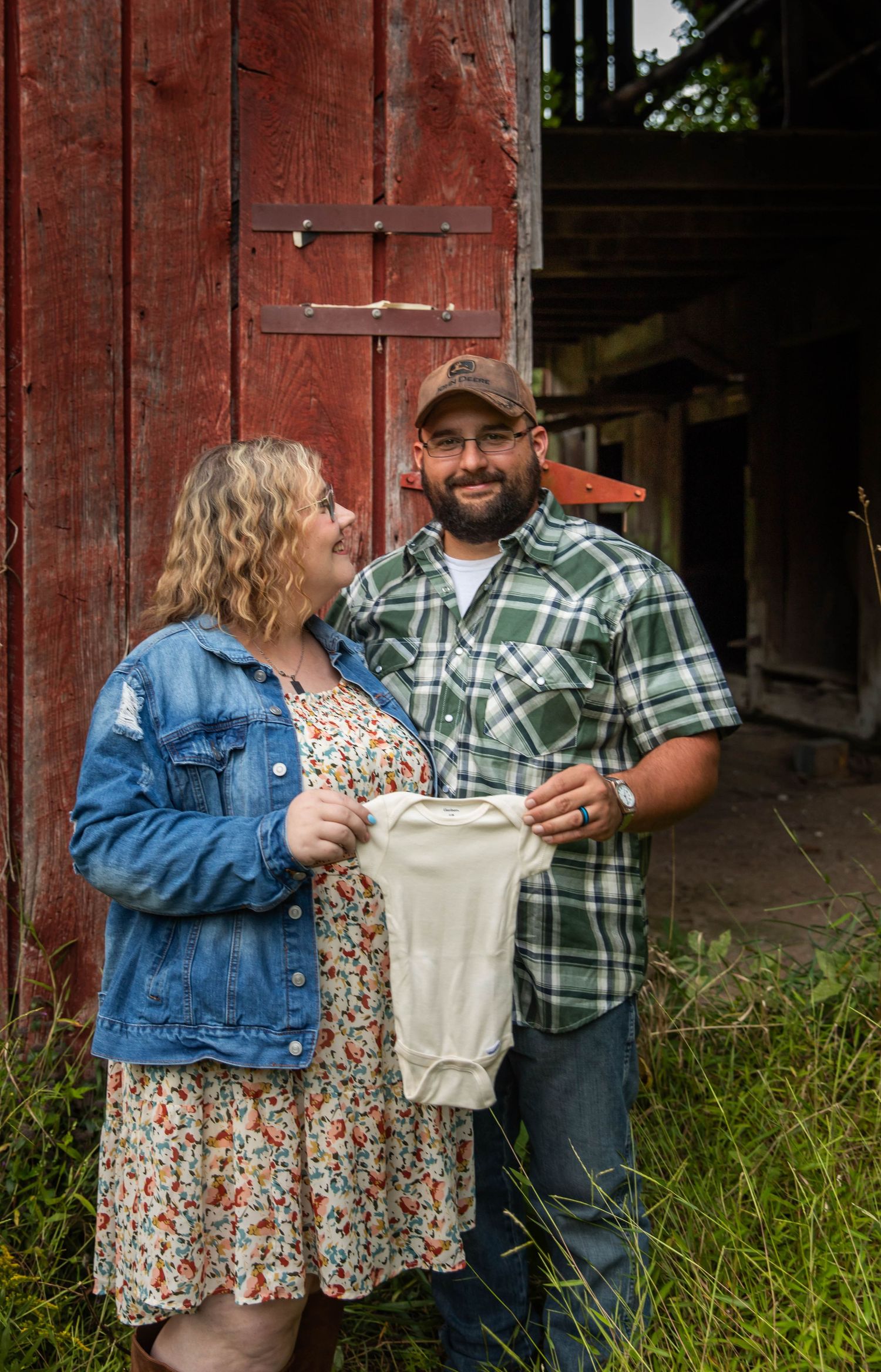 Photo of a couple holding a onesie and looking at one another while standing in front of a red bar for pregnancy announcement photos