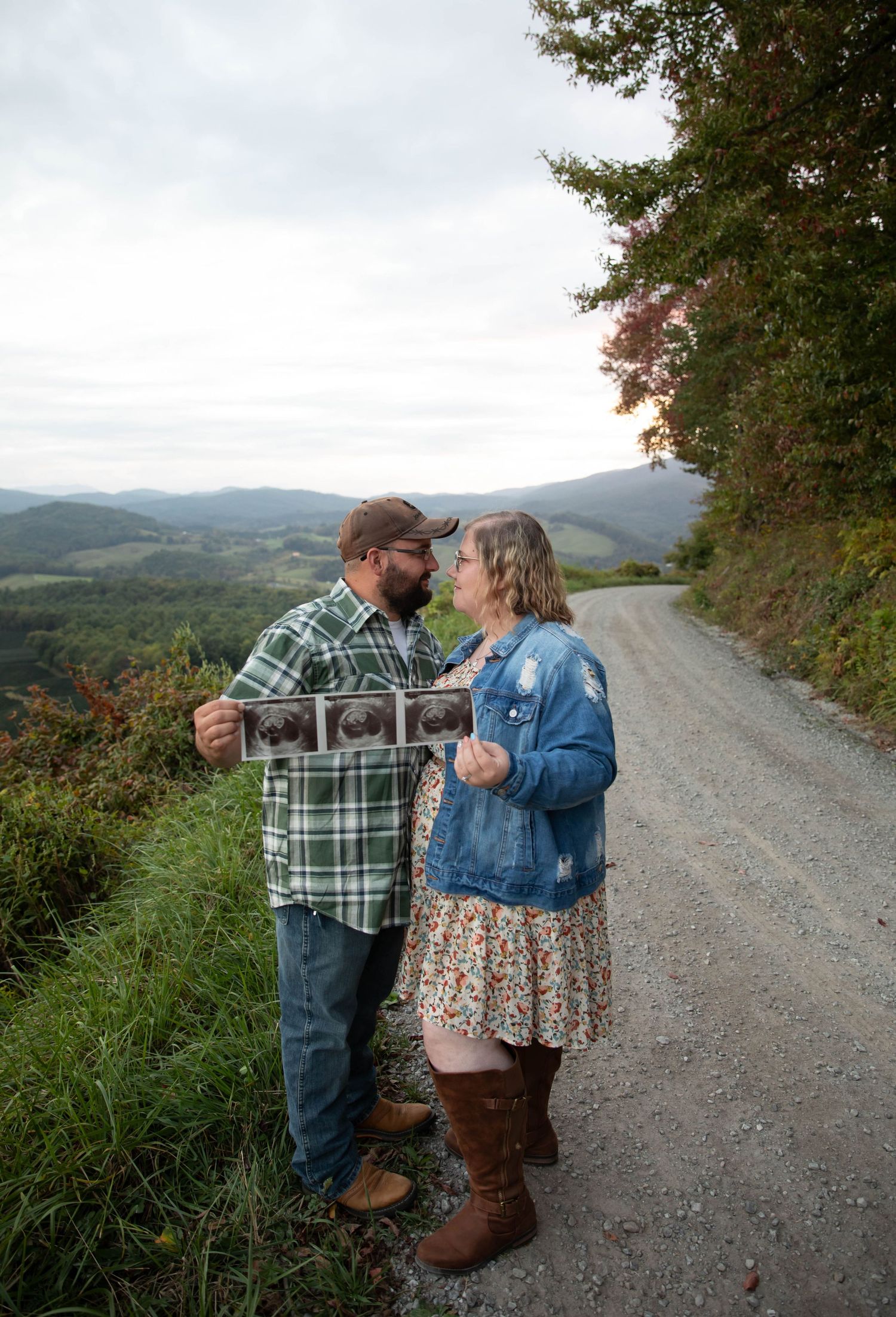 Photo of a couple standing on a mountainside holding ultrasound pictures and looking at one another during a pregnancy announcement shoot