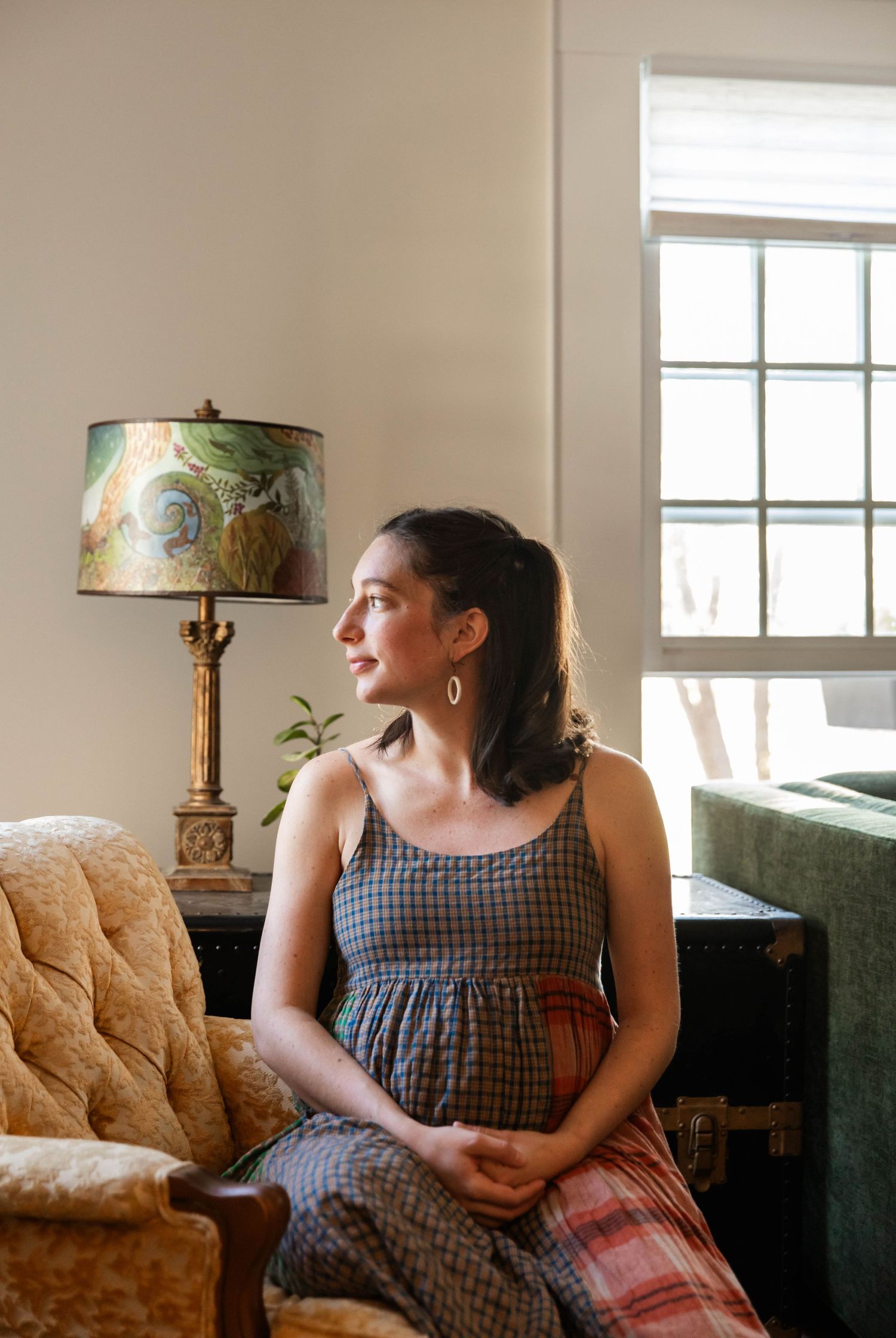 Photo of a pregnant woman sitting in a chair, holding her stomach and looking out the window during a maternity photo shoot