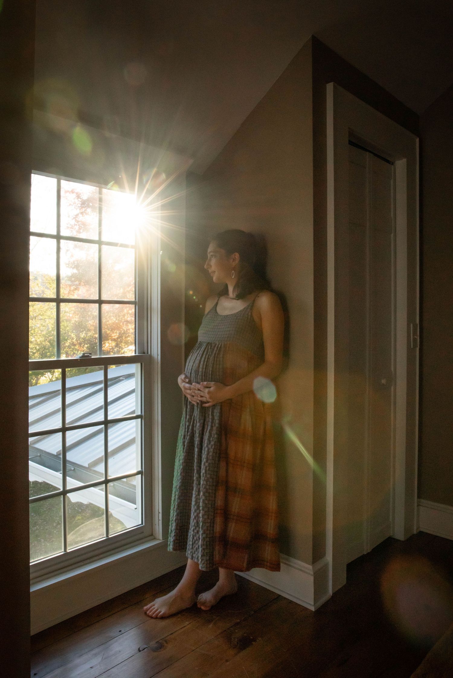 Photo of a pregnant woman standing at a window as light streams in, holding her stomach and staring out of the window. Maternity photo shoot