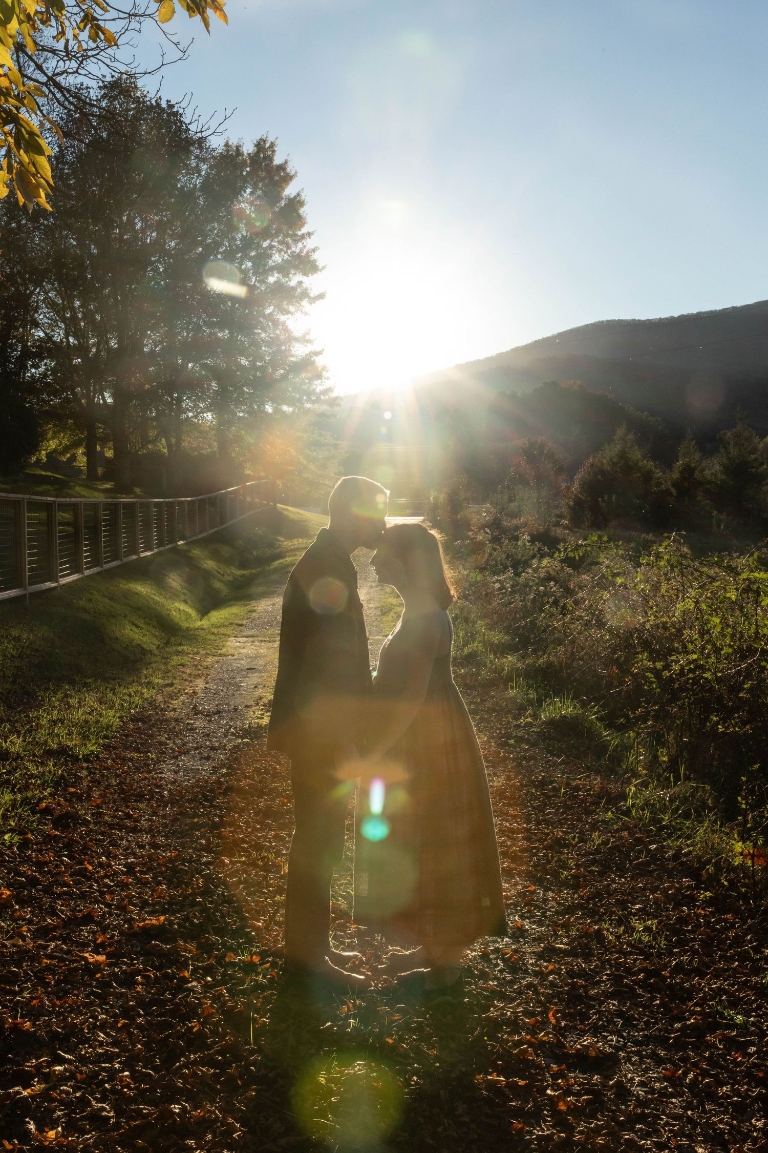 Photo of a couple standing on a trail during sunset. They are facing each other, holding hands, and he is kissing her forehead.