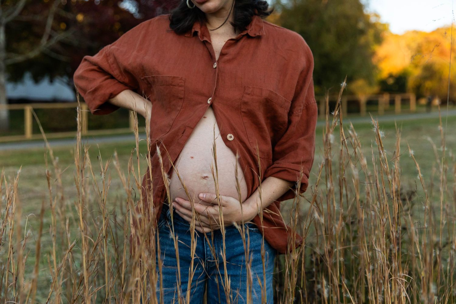 Photo of a pregnant woman standing in a field with tall grass during sunset. Her shirt is partially open, and she is touching her stomach.