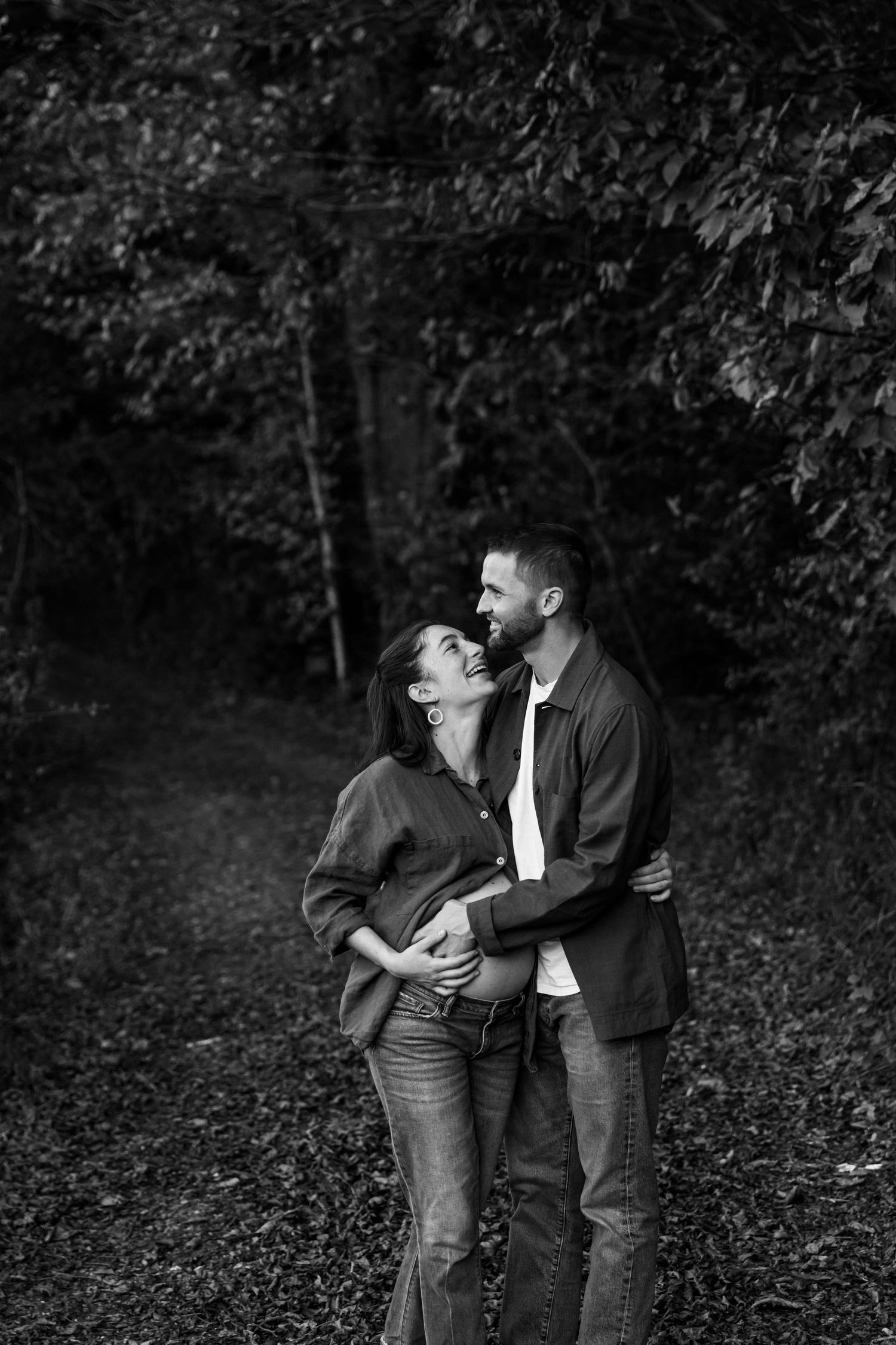 Black and white photo of a couple on a trail in the woods. She is pregnant, and they are both touching her stomach and smiling at each other