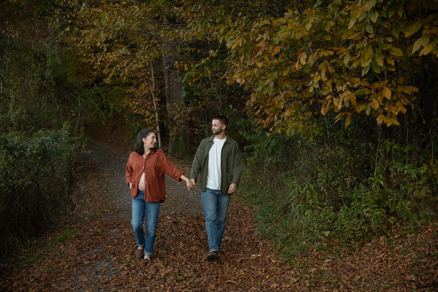 Photo of a couple walking hand in hand through the woods. She is pregnant, and they are smiling at each other