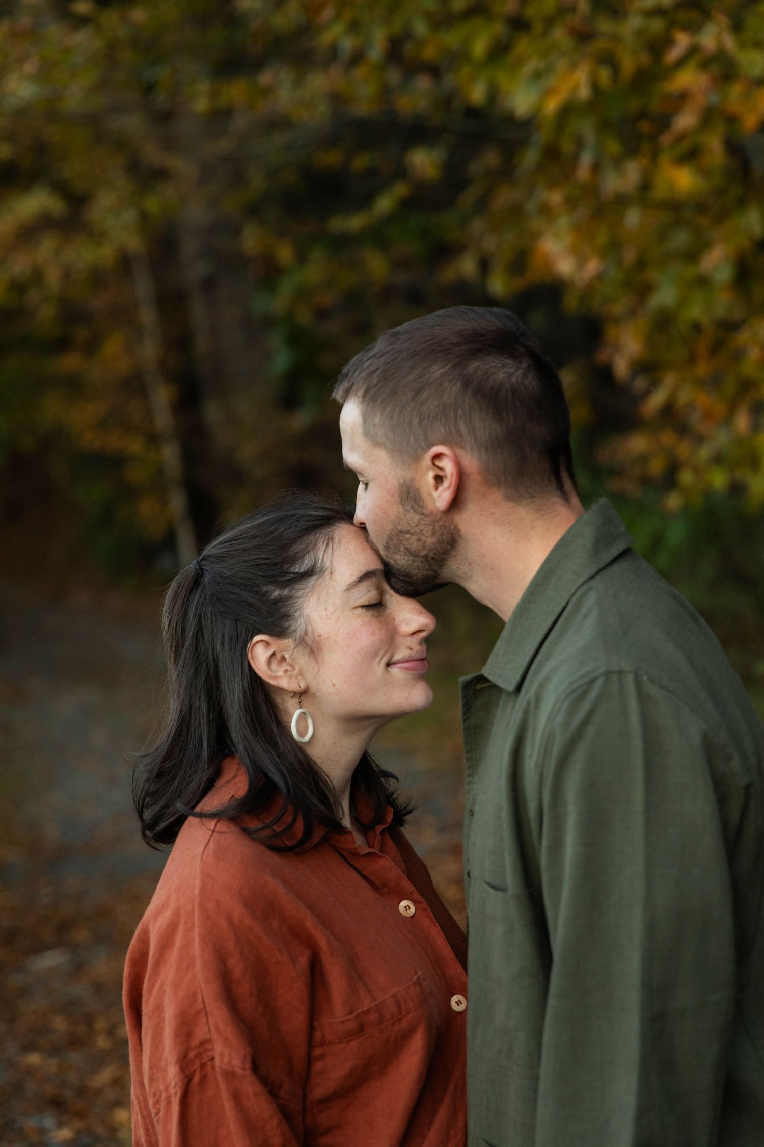 Photo of a couple in the woods. He is kissing her forehead and she is smiling with her eyes closed. Taken during a couples photo shoot