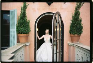 A bride in a classic white ballgown stands in an arched Mediterranean doorway flanked by potted cypress trees.