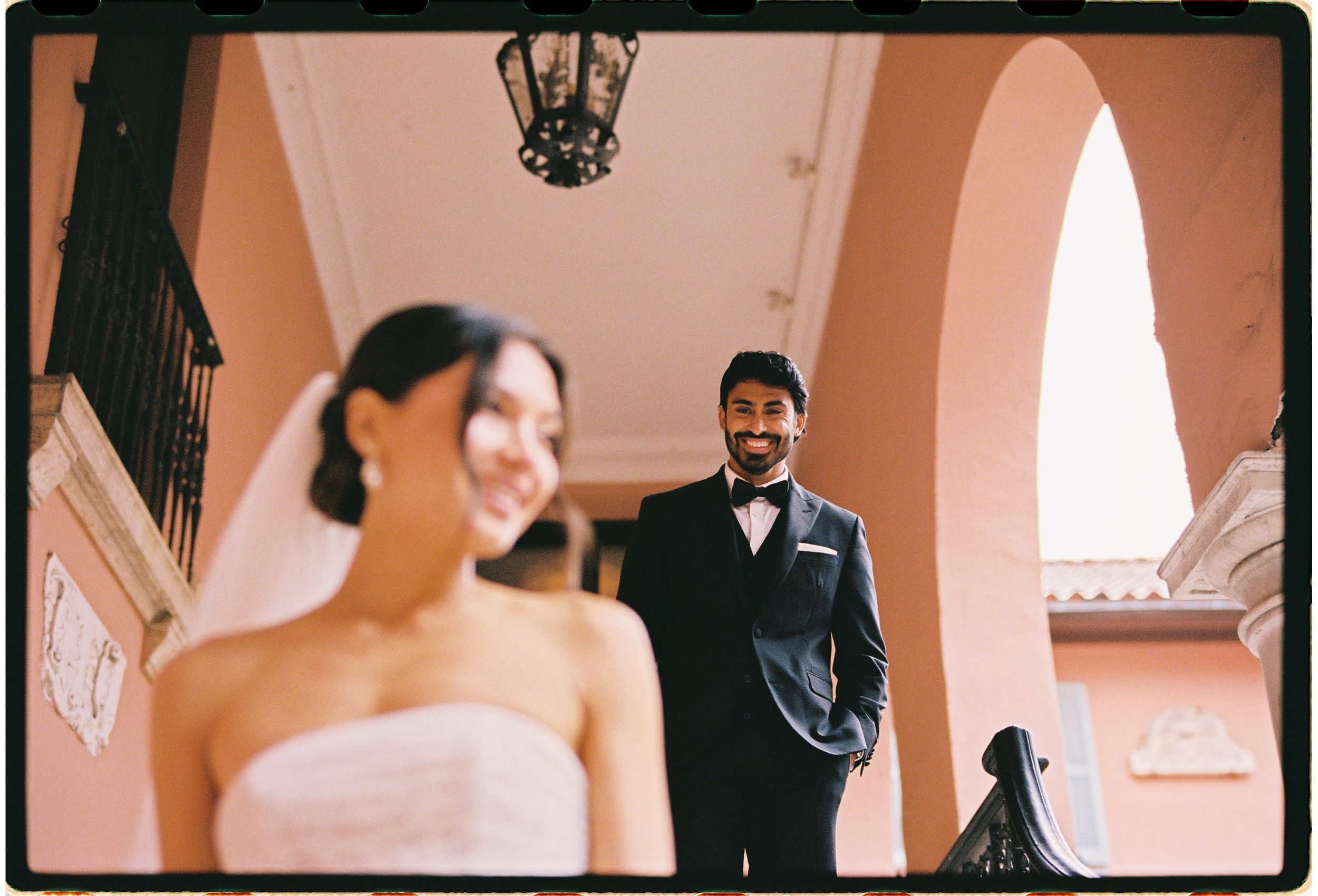 A romantic wedding scene captured on film showing a couple in formal attire under coral-colored arches.