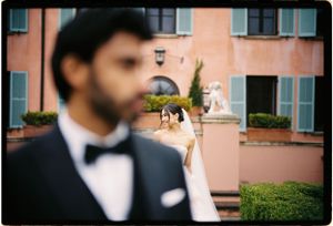 A soft focus wedding photograph showing a groom in a black bowtie with a blurred pink building and hedges in the background.