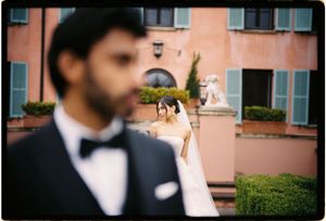 A bride in a white dress stands behind a person wearing a black tuxedo and bowtie at a pink Mediterranean-style venue.