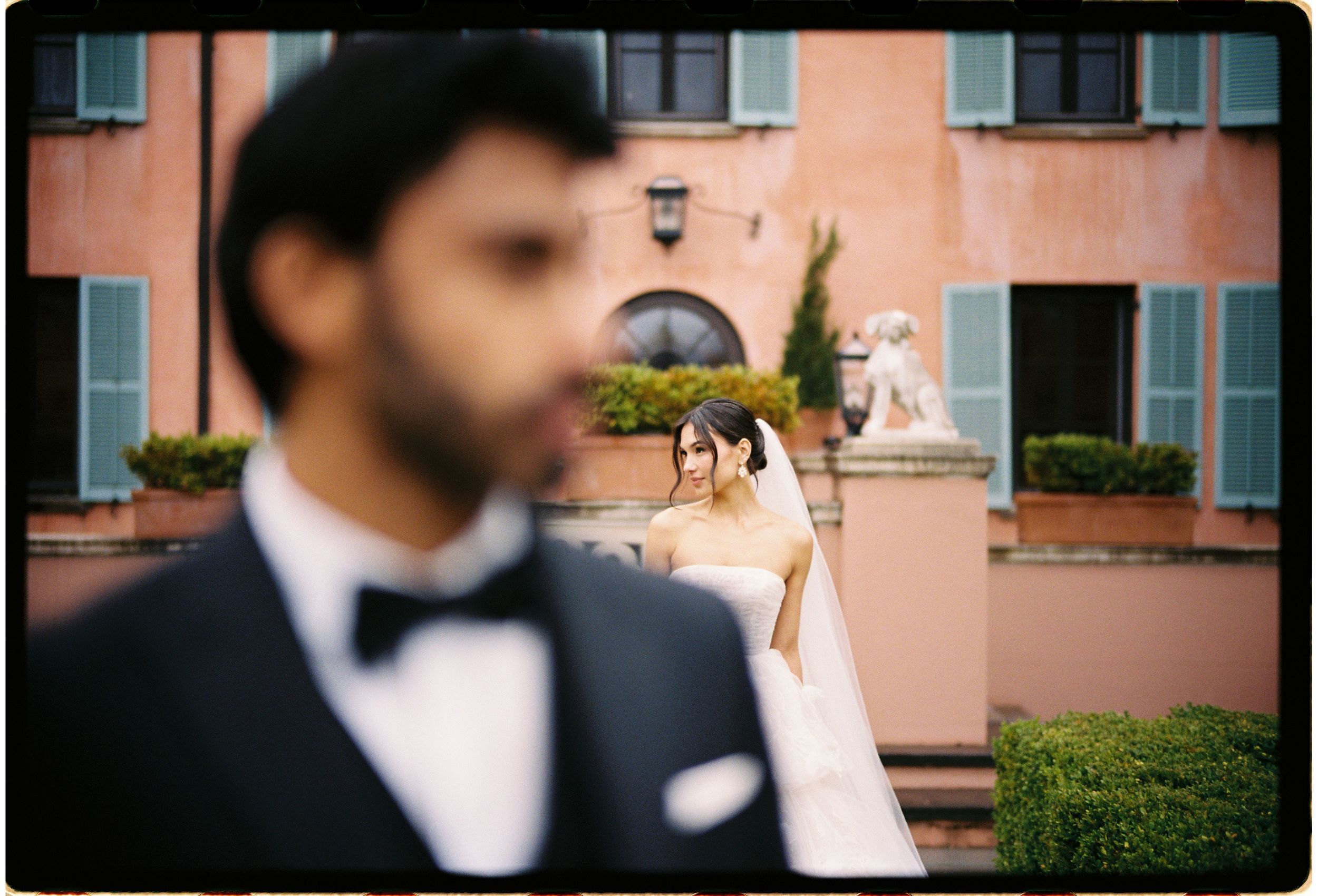 A stylish wedding photo captured at a peach-colored estate building with elegant landscaping and architecture in view.