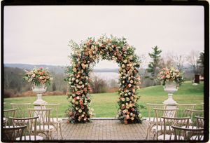An elegant floral wedding arch with pink and white flowers stands on a hilltop overlooking a scenic landscape.