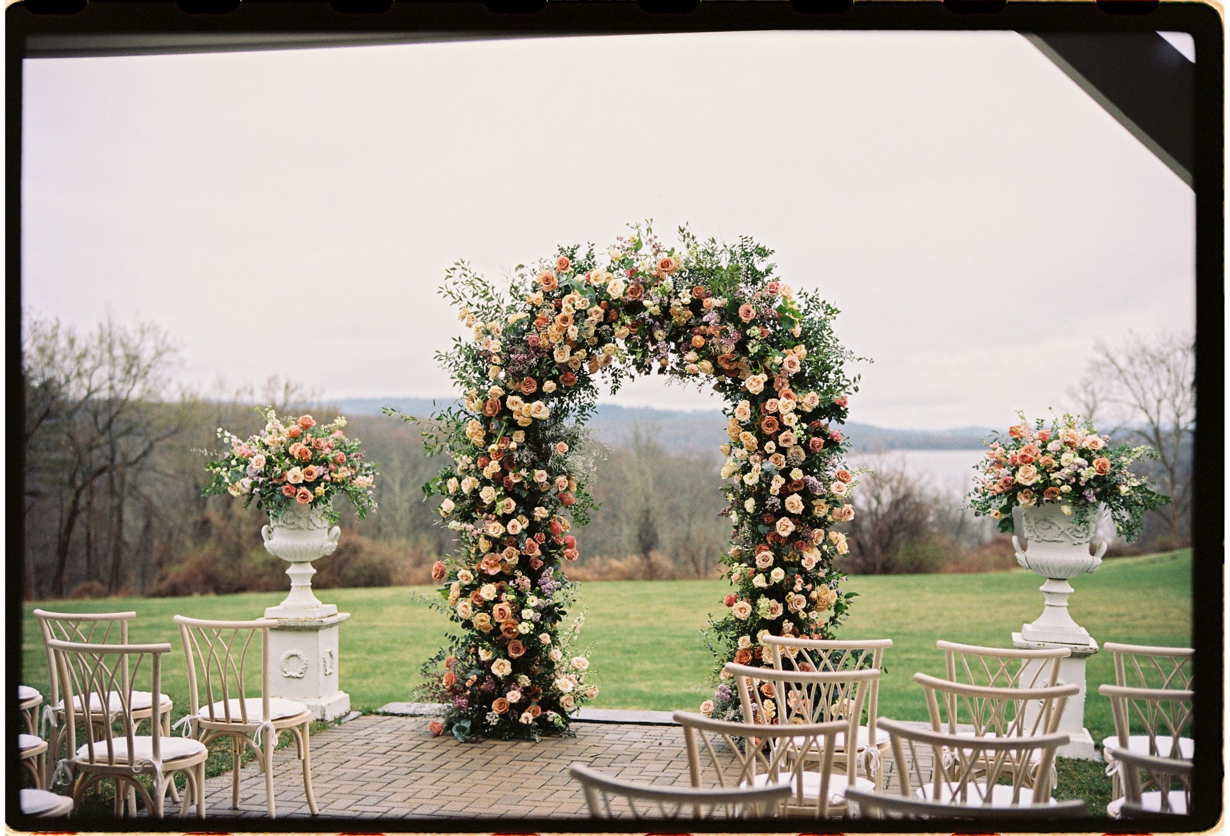 An elegant floral wedding arch with peach and cream blooms overlooks a scenic lake view during an outdoor ceremony setup.