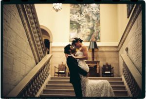 A couple shares a romantic embrace on an elegant staircase with vintage architectural details in warm lighting.