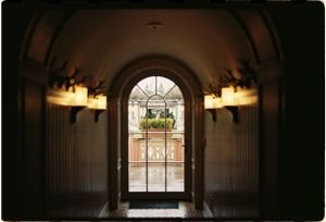 A dimly lit hallway with arched windows and wall sconces leads to a bright window with a wreath.