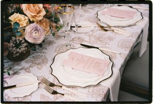 Elegant table setting with pink and gold plates, lace tablecloth, and blush roses in floral arrangement.