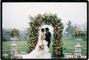 A romantic wedding ceremony under a lush floral arch with peach roses and white lanterns on both sides in a misty garden setting.
