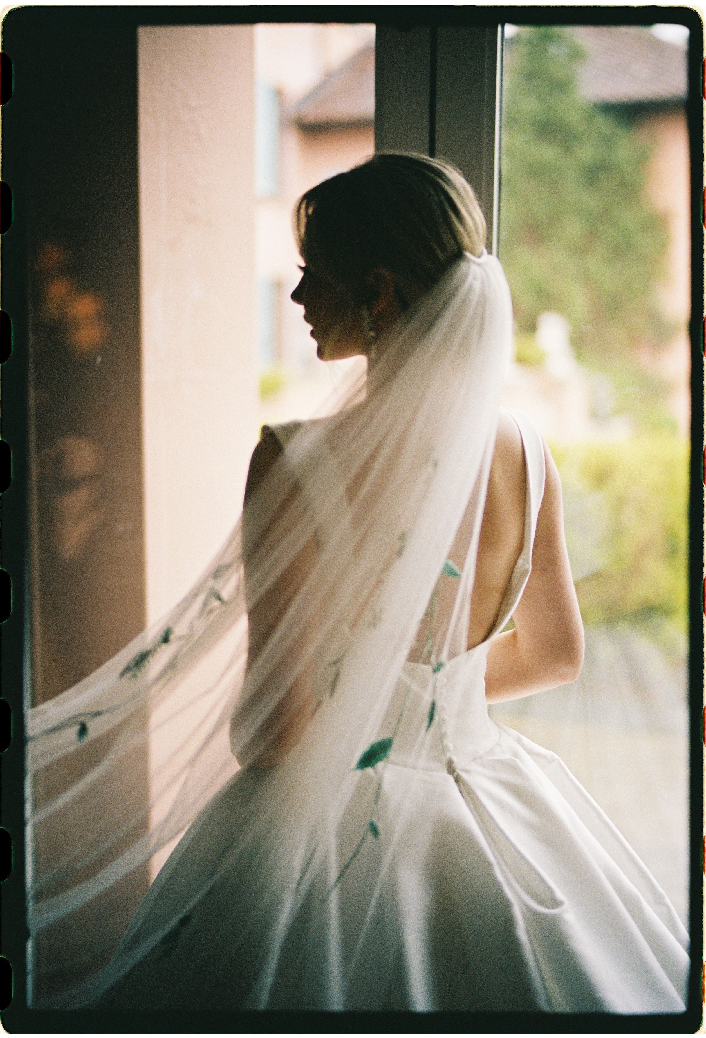 Bride in elegant white wedding dress and veil stands in silhouette by window with flowing fabric catching light.