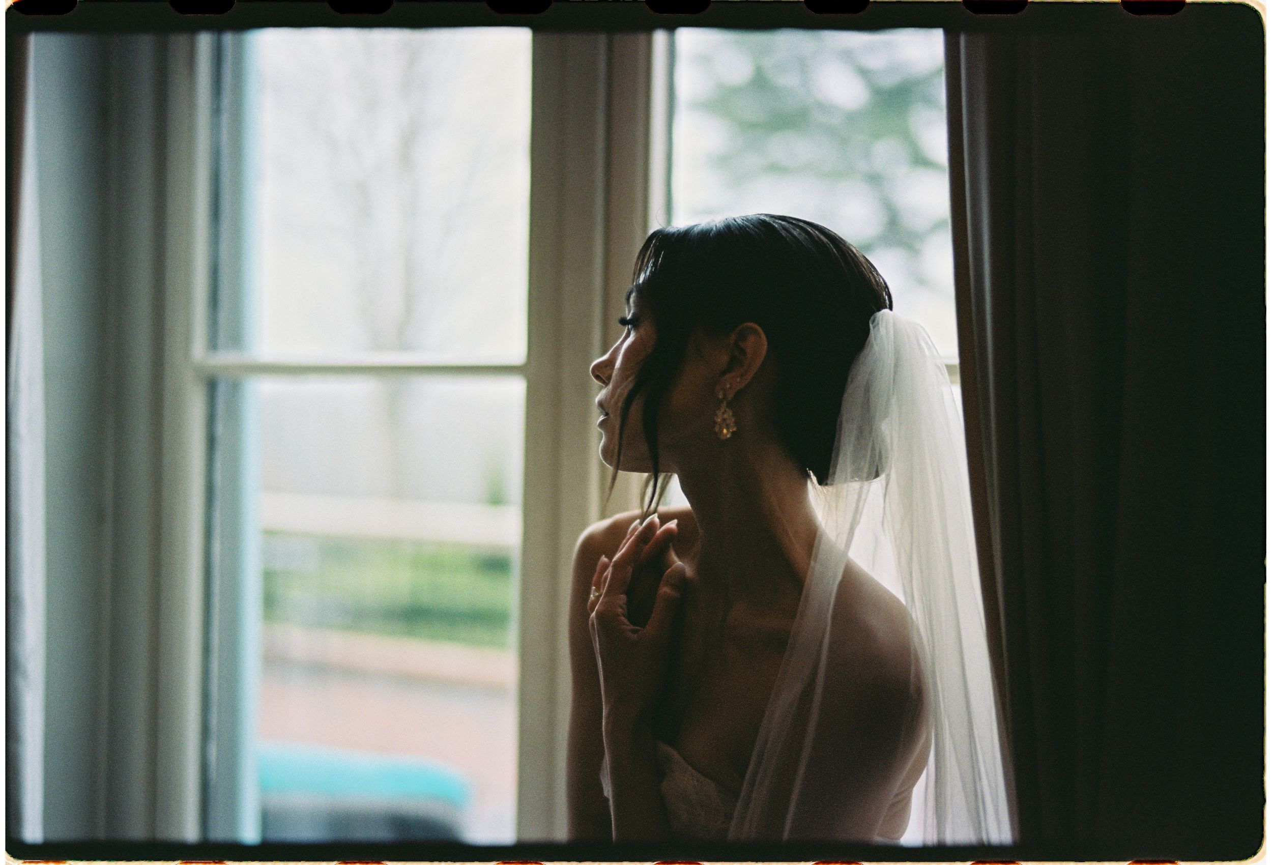 Silhouette of a bride in a veil standing by a window in soft natural light.