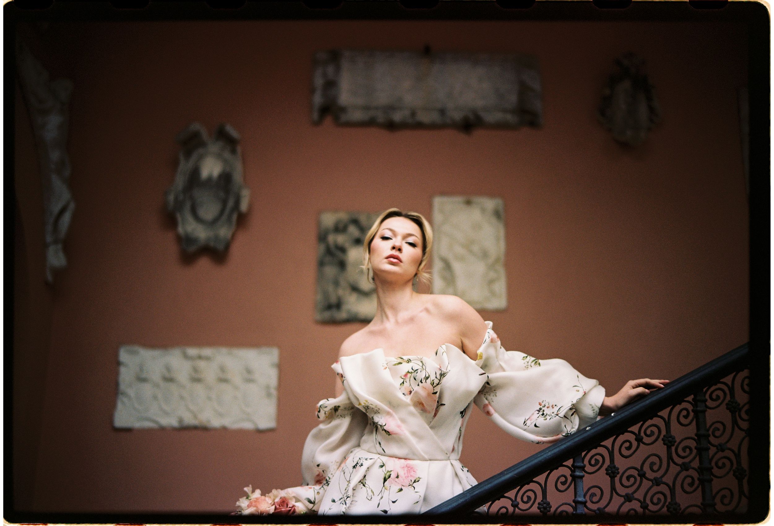 A person in an elegant off-shoulder gown poses dramatically on a staircase with decorative wall art in the background.