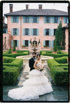 A bride and groom pose for a wedding photo in front of an elegant Italian villa with manicured hedges and fountain.