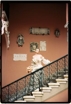 Elegant figure in a vintage gown descends grand staircase with ornate iron railing and classical artwork on terracotta walls.