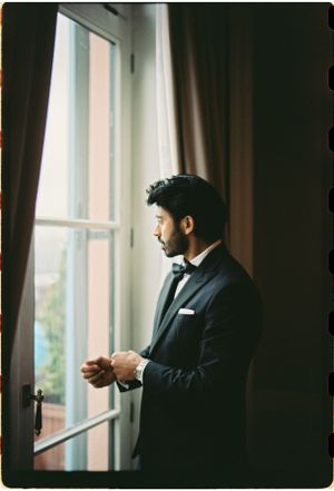 Groom in classic black tuxedo looking pensively out window in dramatic lighting.