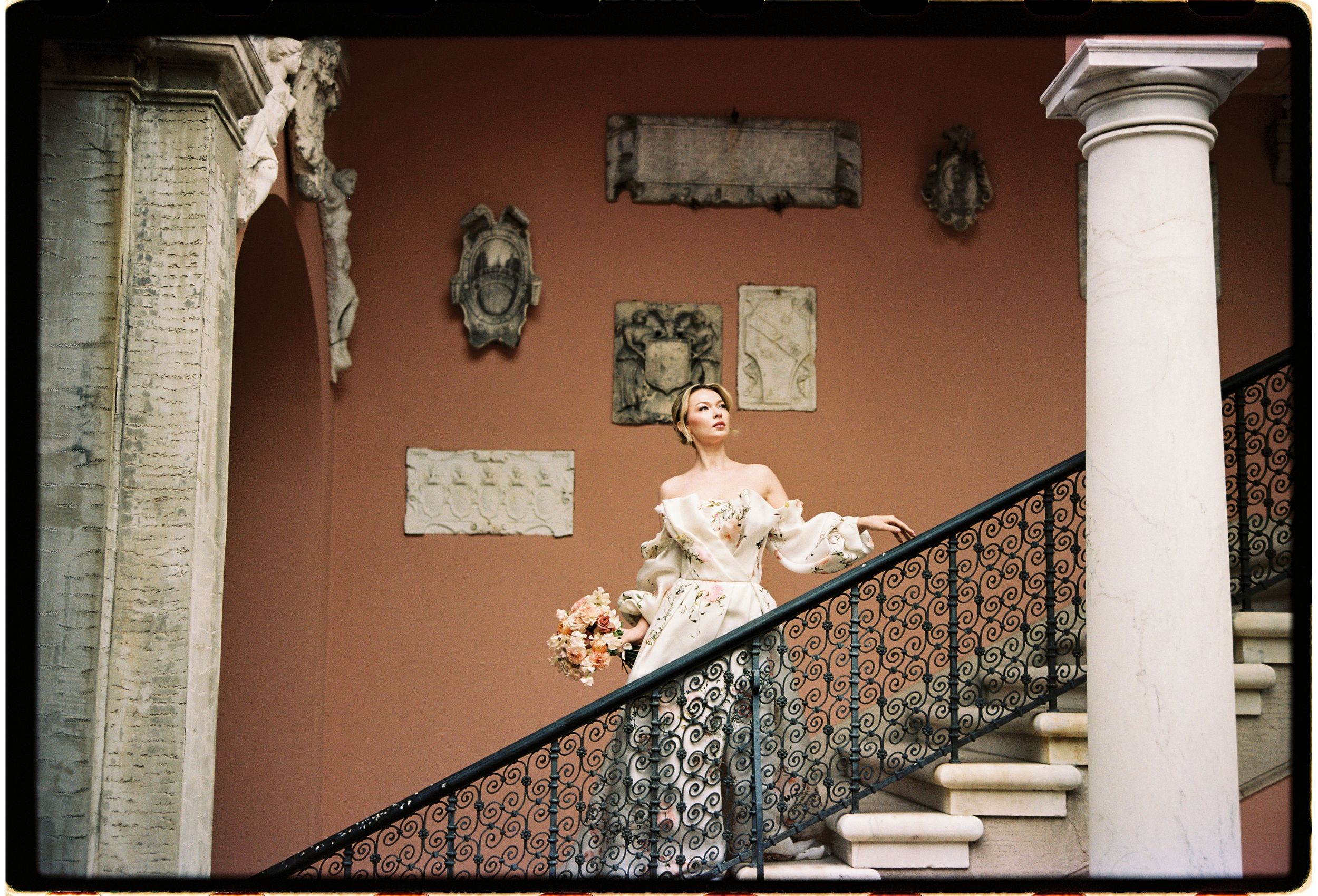 An elegant staircase with ornate iron railings and columns leads up against a terra cotta colored wall with decorative accents.