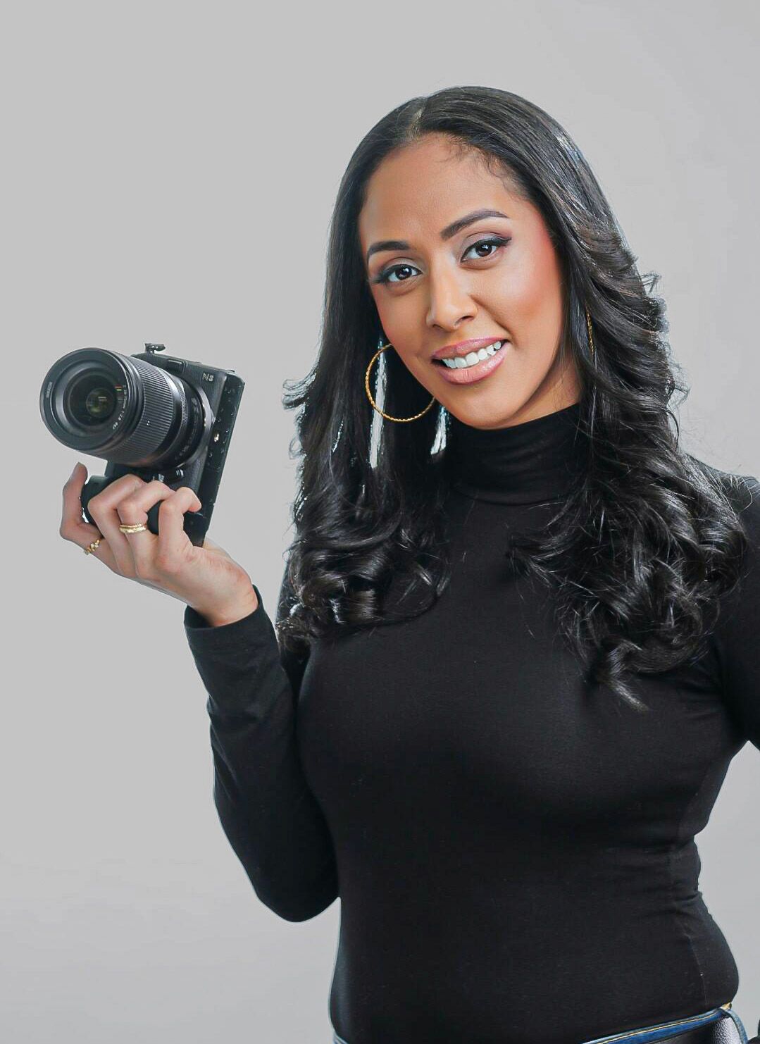 Photographer in black turtleneck displaying camera handling poses against white backdrop.
