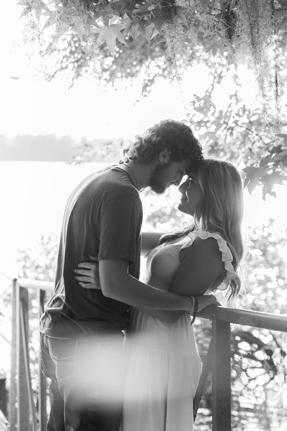 Black and white photo of a romantic couple embracing and leaning in for a kiss on a wooden deck near a lake.