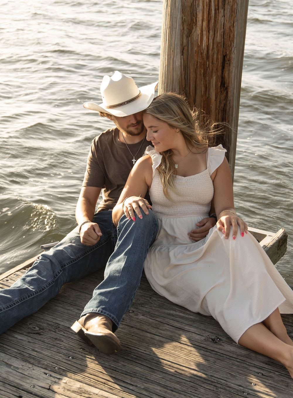 Couple sitting on a wooden dock by the water, man in cowboy hat and jeans, woman in white dress, sharing a romantic moment.