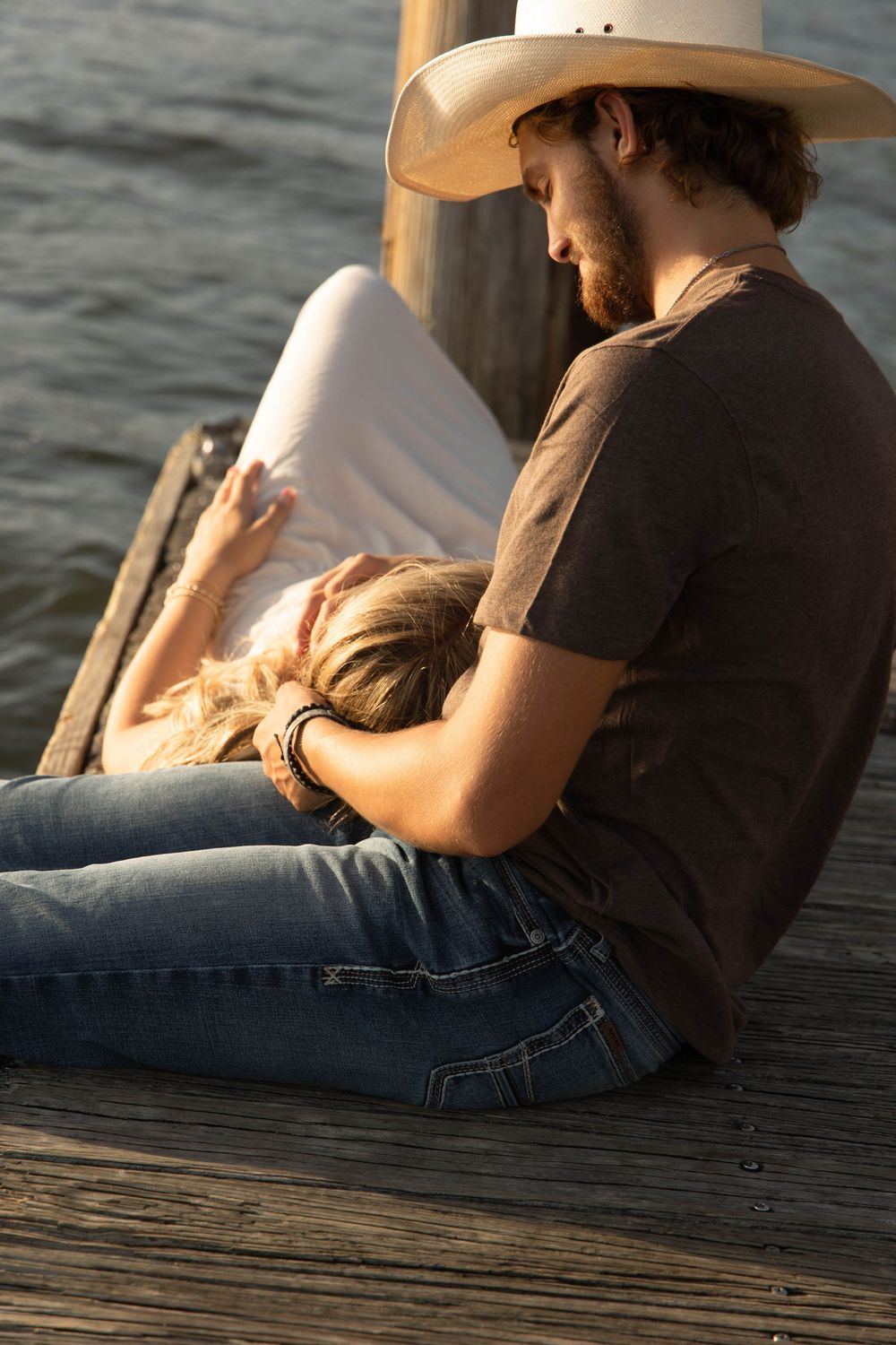 A bearded man in a cowboy hat sits on a wooden dock holding a woman resting her head on his lap.