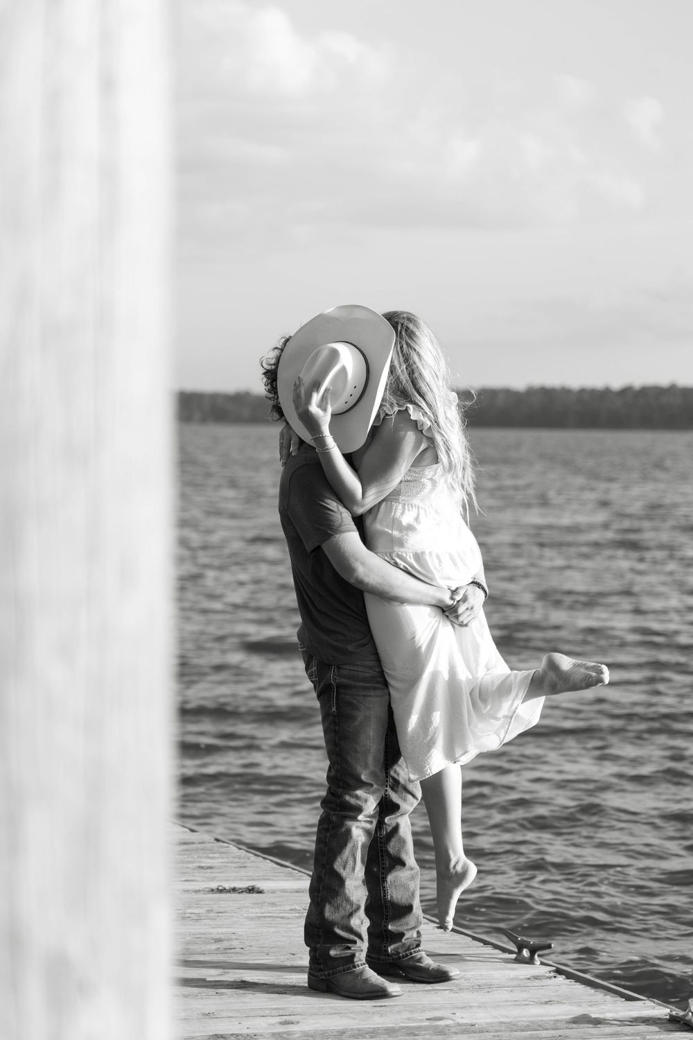 Black and white photo of a couple kissing on a dock by a lake, woman lifted off ground, man wearing cowboy hat.