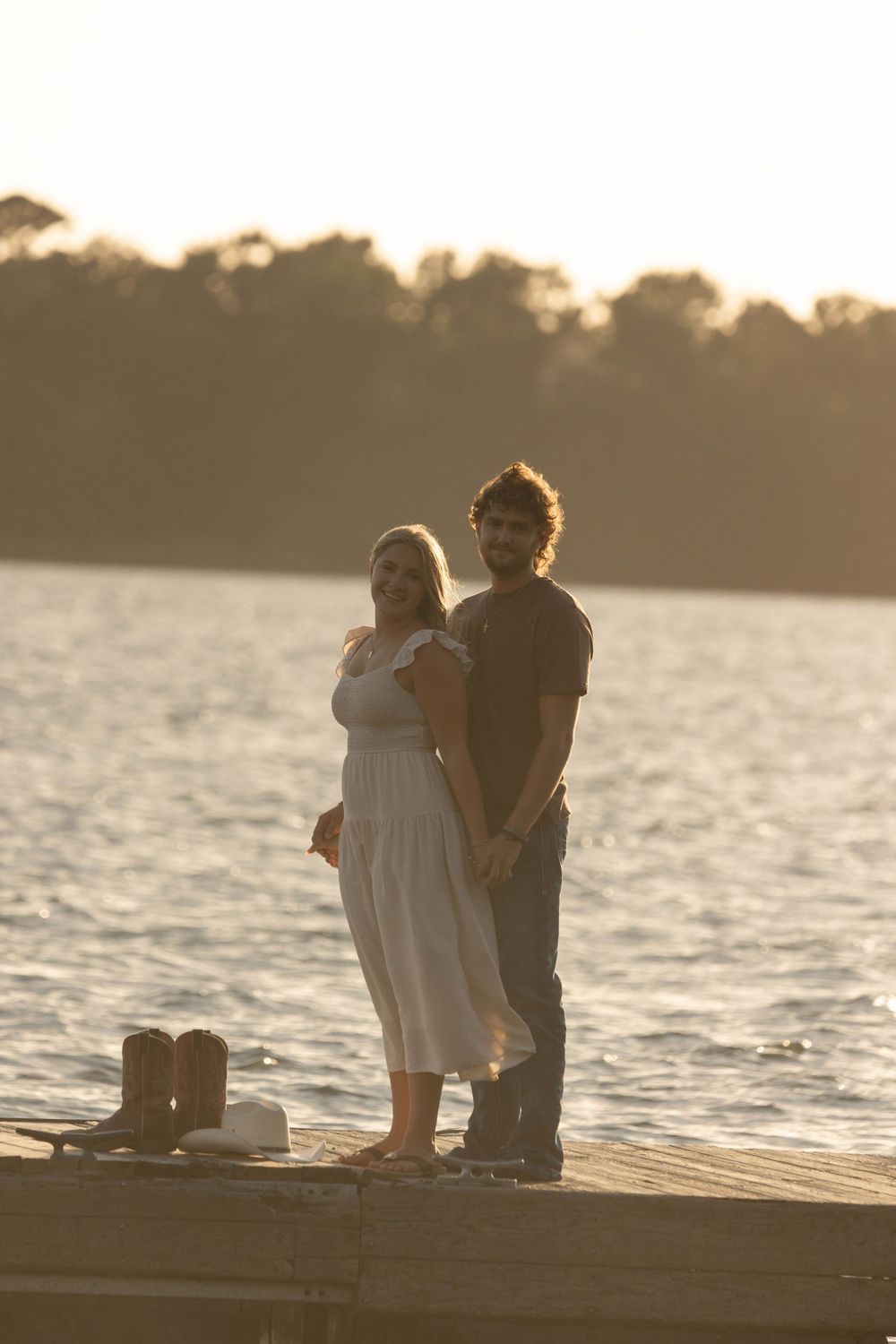 Couple standing on a dock at sunset by a lake, woman in white dress, man in dark shirt, boots nearby.