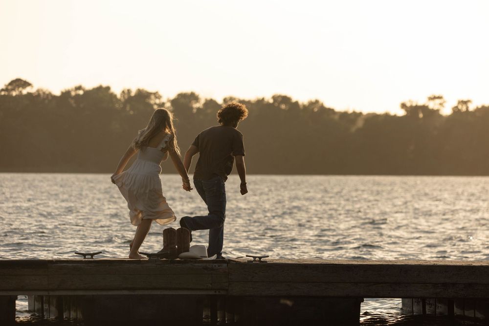 Couple standing on a wooden dock at sunset by a serene lake surrounded by lush green trees.