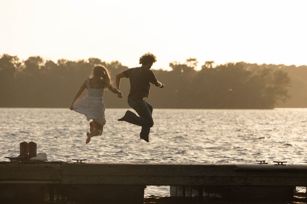 Two people holding hands jumping off a wooden dock into a lake at golden hour sunset.