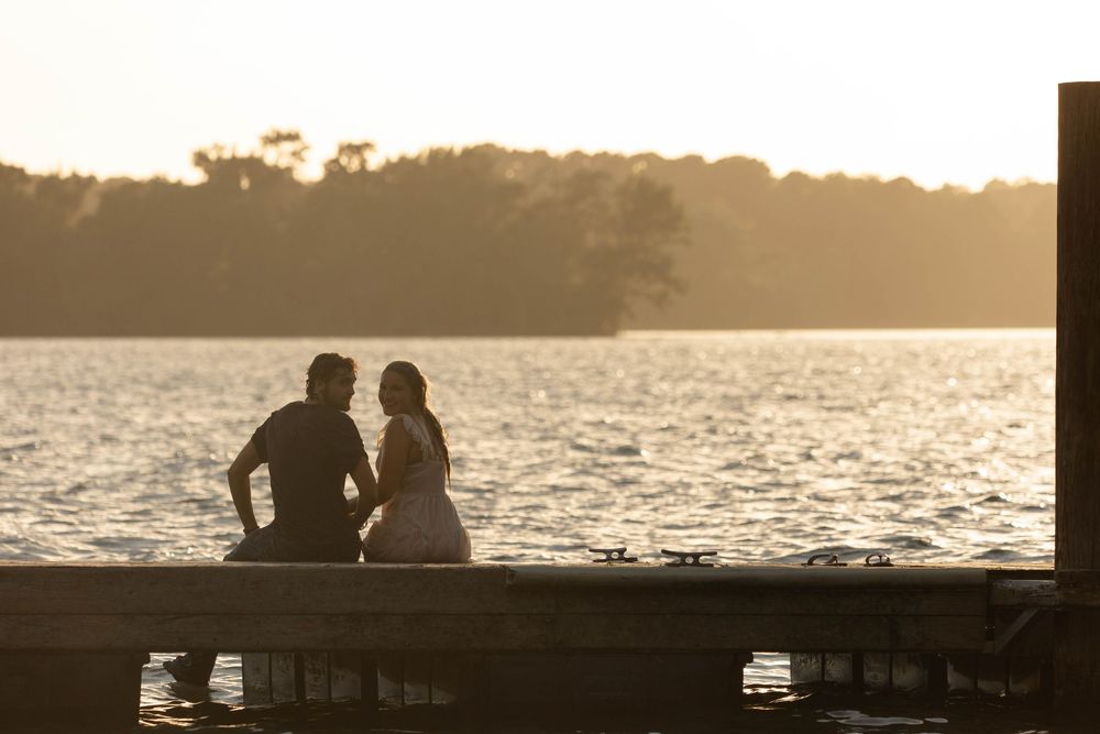 Couple sitting together on a dock at sunset, silhouetted against a golden lake with trees in the background.
