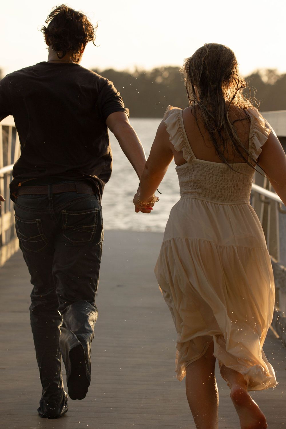 Couple holding hands running on a wooden pier near water at sunset, woman in white dress and man in black shirt.