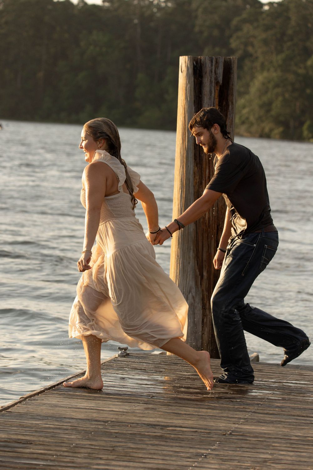 Couple holding hands running barefoot on a wooden dock by a lake at sunset, woman in flowing dress.