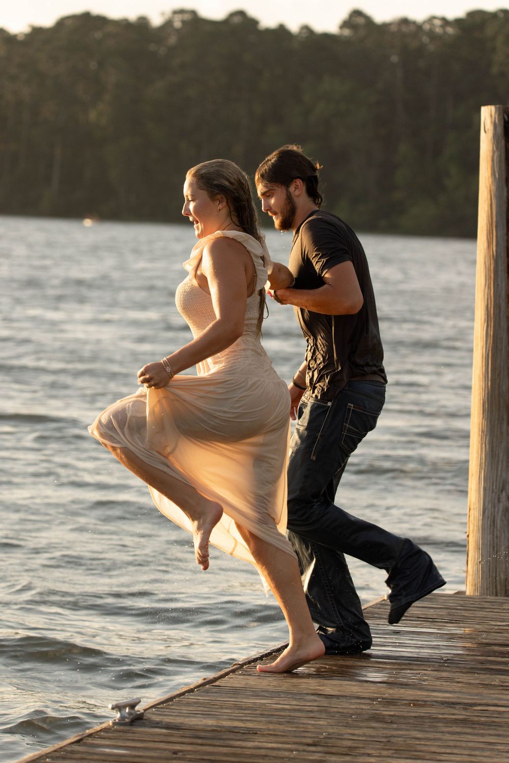 Couple dancing joyfully on a wooden dock by the water at sunset, woman in flowing dress, man in black shirt.