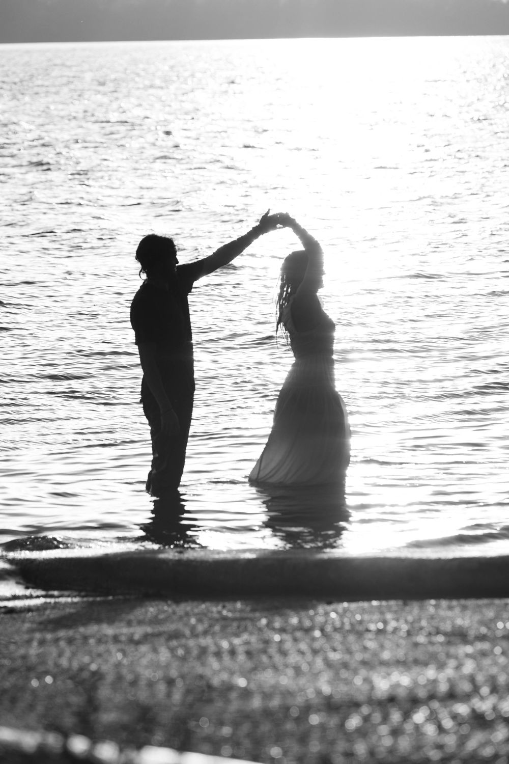 Black and white photo of a couple dancing in the ocean waves at sunset, silhouetted against shimmering water.