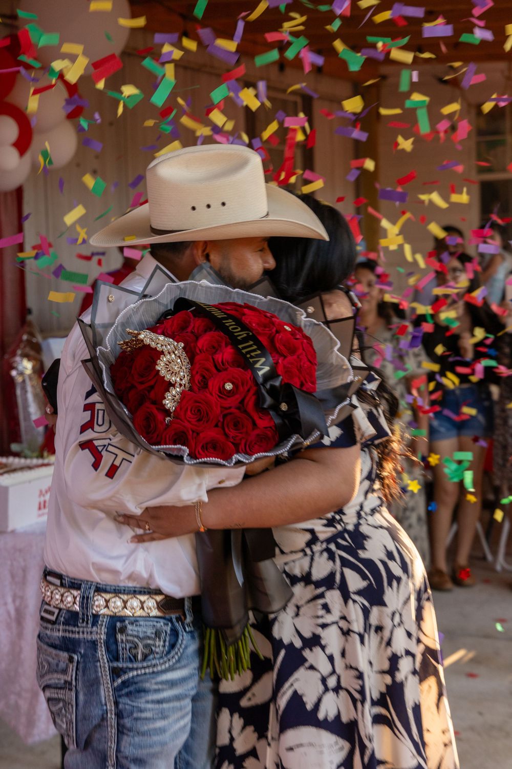 A romantic embrace under colorful confetti with a large red bouquet between them.