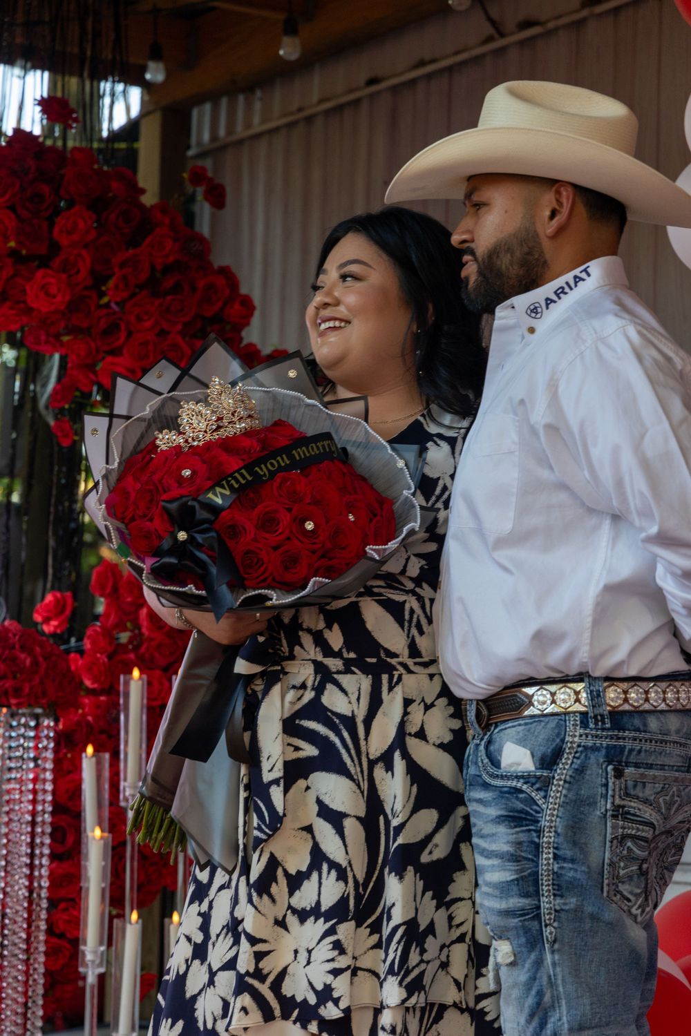 A couple stands together amid red roses holding a beautiful bouquet.