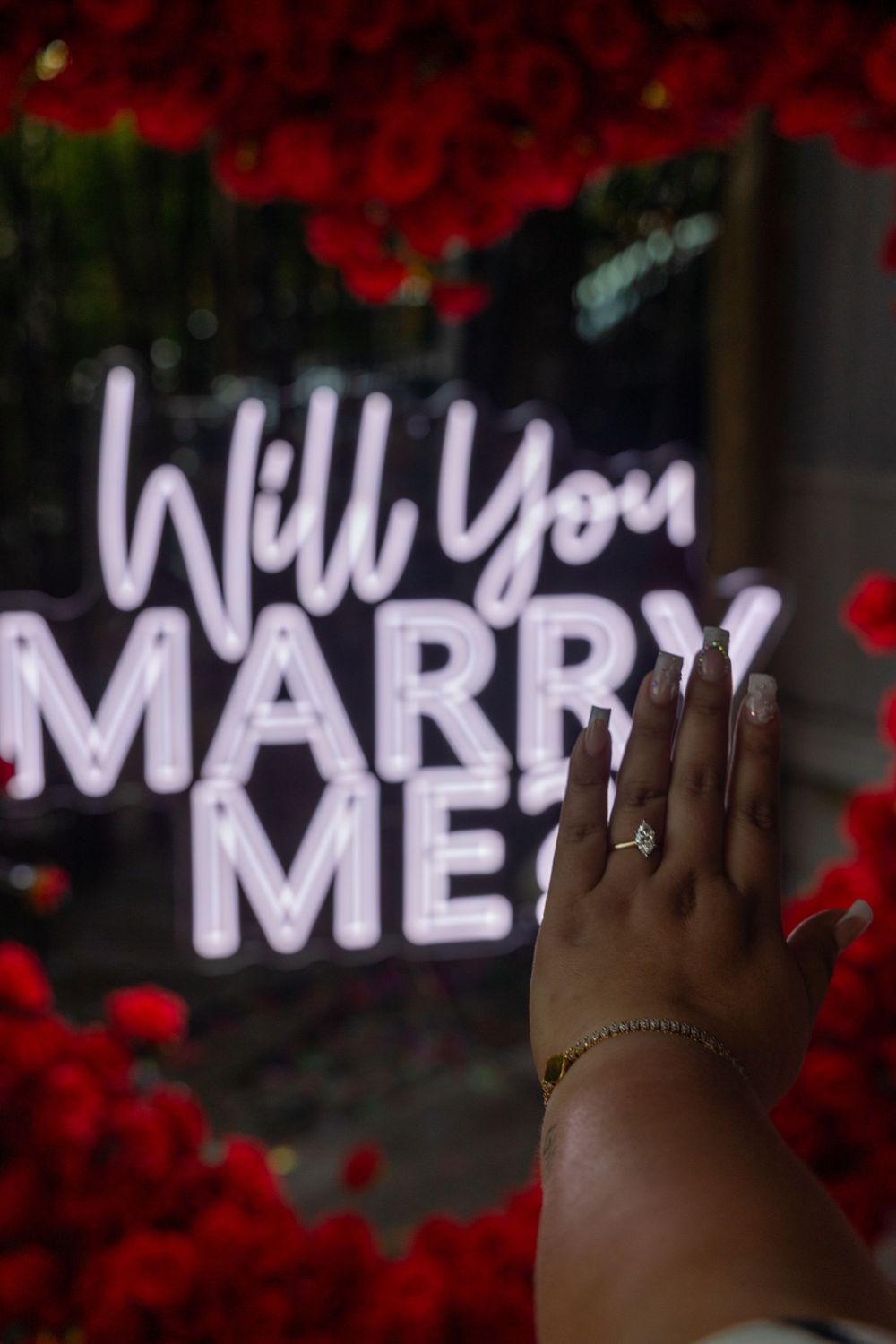 Hand with engagement ring shown against illuminated marriage proposal sign surrounded by red roses.
