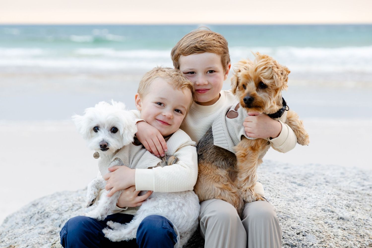 Two siblings sitting on a beach with their dogs hugging and smiling together by the ocean.