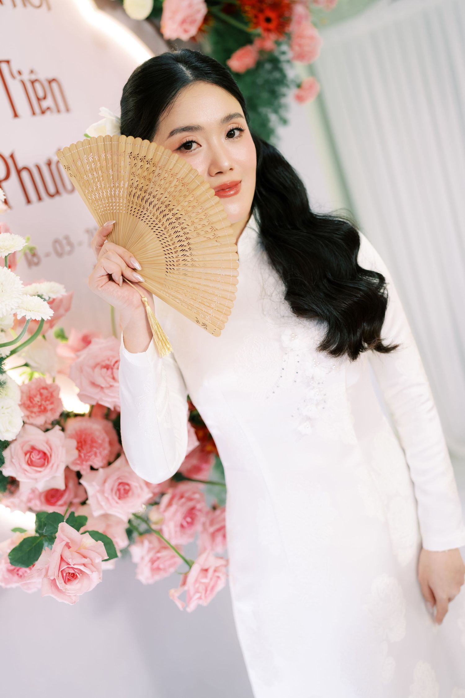 Someone in a white traditional dress holds a wooden fan while posing against a floral backdrop with pink roses.