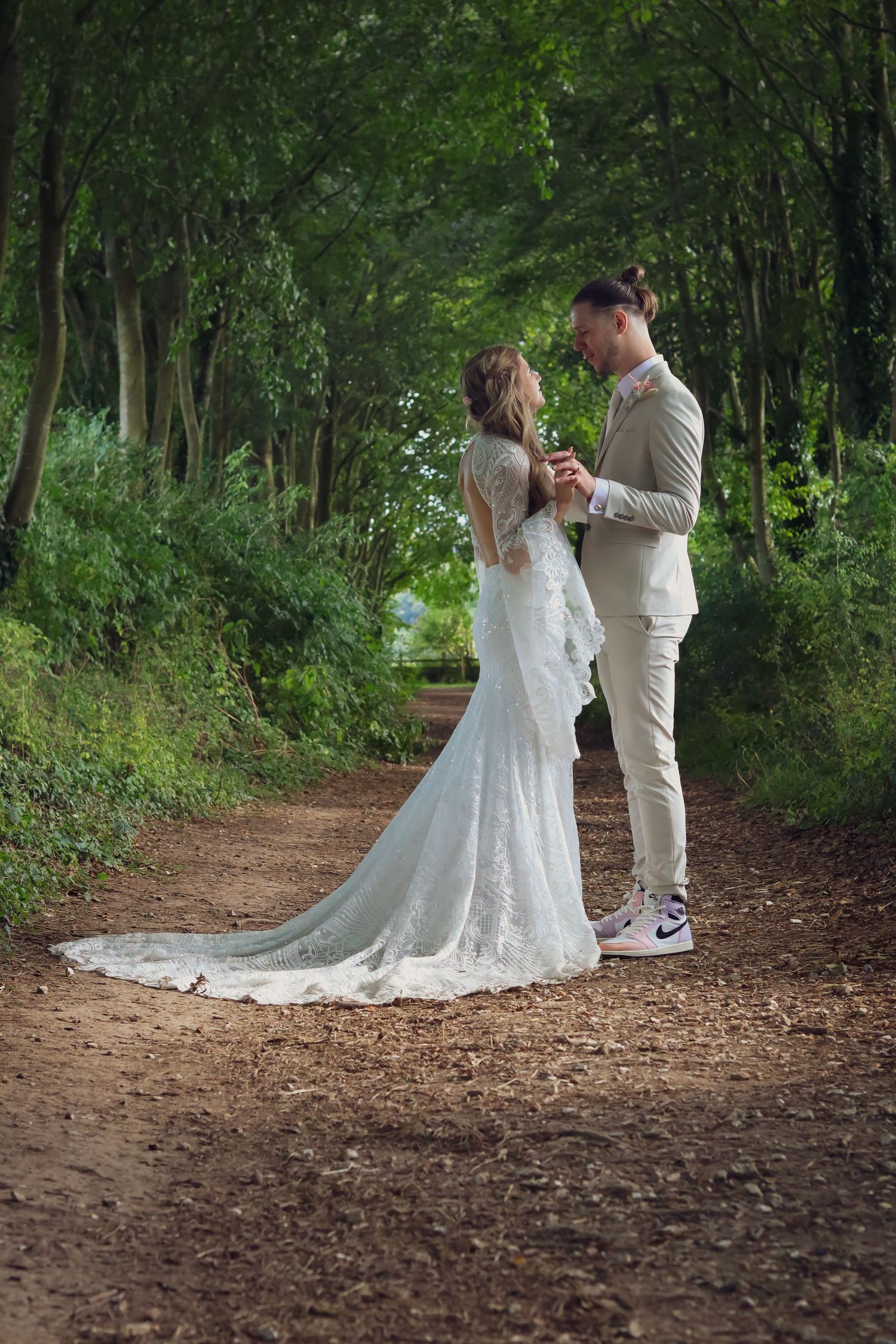 A romantic woodland scene shows a couple in wedding attire sharing a tender moment on a tree-lined dirt path.