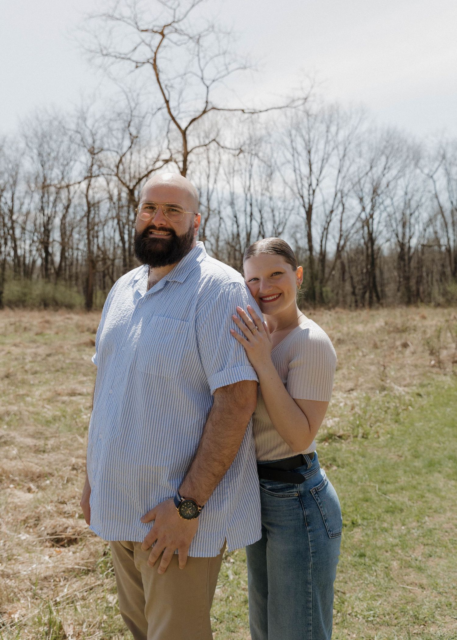 A couple stands together in a sunny field with bare trees in the background on a spring day.